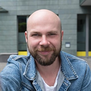 Foto van Gregory van Trusted Shops. Hij heeft een kaal hoofd, een bruine baard en snor. Hij heeft een denim shirt aan met een wit t-shirt eronder.