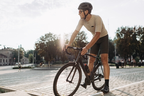 Sportieve jongeman met actieve kleding, helm en bril, zittend op een zwarte fiets in de buitenlucht.